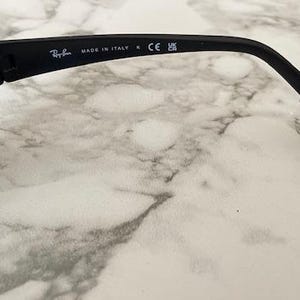 May include: Black sunglasses featuring a matte finish frame. The inner temple arm displays the white printed text Ray-Ban, MADE IN ITALY, the CE mark, and the UKCA symbol. The design includes a curved temple tip and a dark tinted lens.