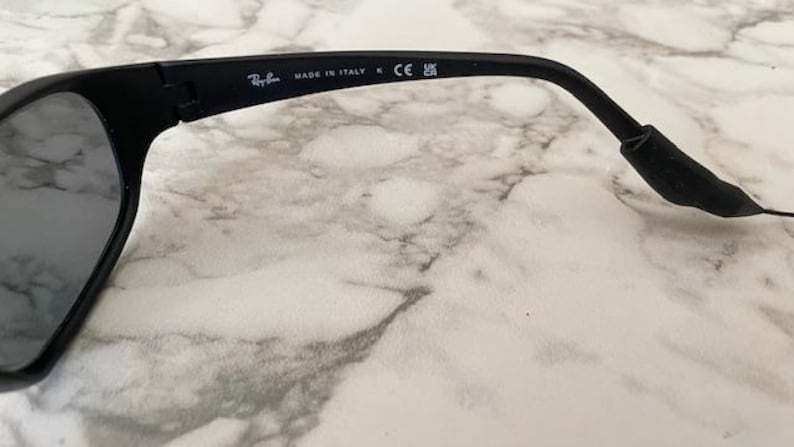 May include: Black sunglasses featuring a matte finish frame. The inner temple arm displays the white printed text Ray-Ban, MADE IN ITALY, the CE mark, and the UKCA symbol. The design includes a curved temple tip and a dark tinted lens.
