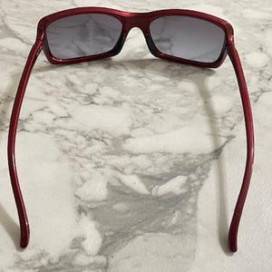 May include: Red-framed sunglasses with gray lenses. The sunglasses have a rectangular shape and a glossy finish. The temples are thin and curve gently towards the ear. The sunglasses are resting on a marble surface.