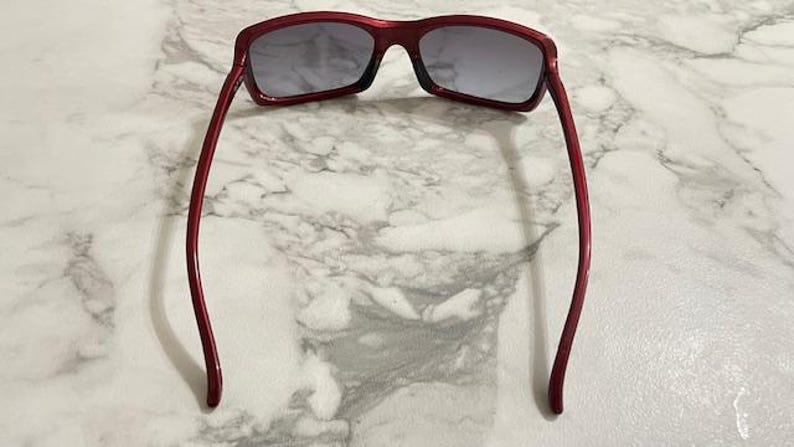 May include: Red-framed sunglasses with gray lenses. The sunglasses have a rectangular shape and a glossy finish. The temples are thin and curve gently towards the ear. The sunglasses are resting on a marble surface.