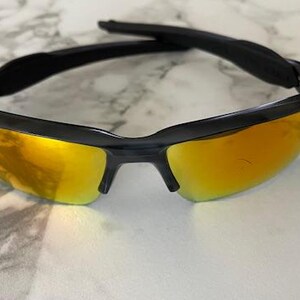 Puede incluir: Gafas de sol negras con lentes doradas. Las gafas tienen un dise&ntilde;o deportivo con montura negra y barra superior curvada. Las lentes son de color dorado brillante y reflectante, sobre una superficie de m&aacute;rmol.