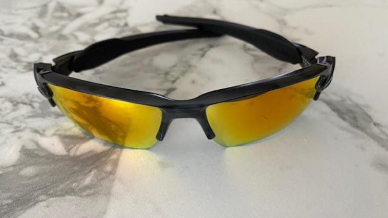 Puede incluir: Gafas de sol negras con lentes doradas. Las gafas tienen un dise&ntilde;o deportivo con montura negra y barra superior curvada. Las lentes son de color dorado brillante y reflectante, sobre una superficie de m&aacute;rmol.