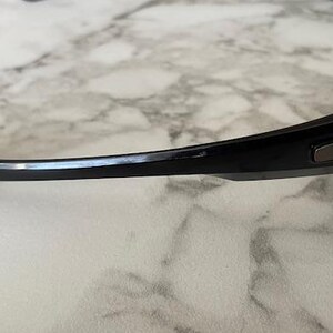 K&ouml;nnte beinhalten: Schwarze Sonnenbrille mit gebogenem Rahmen. Die Sonnenbrille hat ein schlankes, modernes Design mit dunklen Gl&auml;sern und einem schwarzen Rahmen. Das Oakley-Logo ist auf dem B&uuml;gel sichtbar.