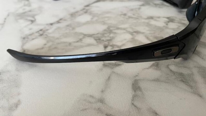 K&ouml;nnte beinhalten: Schwarze Sonnenbrille mit gebogenem Rahmen. Die Sonnenbrille hat ein schlankes, modernes Design mit dunklen Gl&auml;sern und einem schwarzen Rahmen. Das Oakley-Logo ist auf dem B&uuml;gel sichtbar.