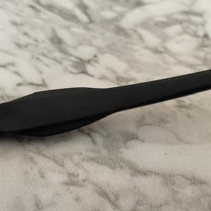 Peut inclure: Gros plan sur une branche de monture de lunettes noire. La branche est faite d'un mat&eacute;riau lisse et mat. La monture repose sur une surface &agrave; motif marbr&eacute; blanc et gris. La branche a un design incurv&eacute; pour un ajustement confortable.