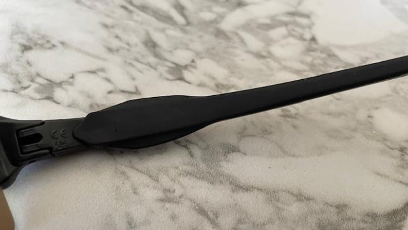 Peut inclure: Gros plan sur une branche de monture de lunettes noire. La branche est faite d'un mat&eacute;riau lisse et mat. La monture repose sur une surface &agrave; motif marbr&eacute; blanc et gris. La branche a un design incurv&eacute; pour un ajustement confortable.
