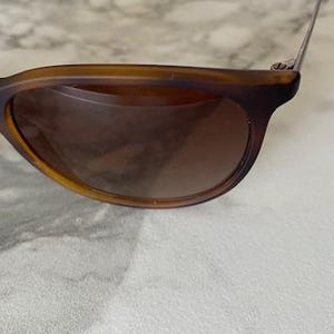May include: Brown sunglasses with a classic design. The frames are a warm brown color, and the lenses are a darker shade of brown. The sunglasses are resting on a white surface.