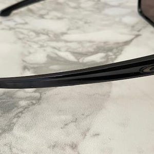 May include: Black sunglasses with a sleek, modern design. The frames are glossy black with a subtle stripe detail and the Oakley logo. The lenses are dark, providing shade. The sunglasses are resting on a marble-patterned surface.