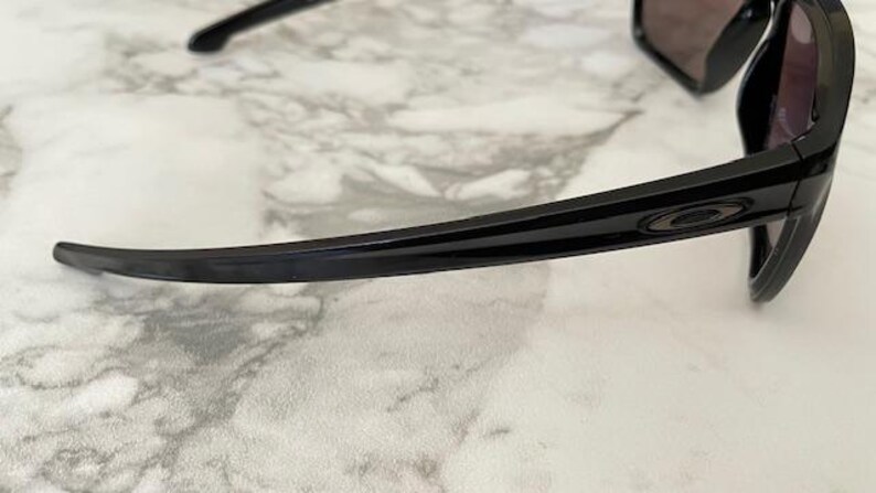 May include: Black sunglasses with a sleek, modern design. The frames are glossy black with a subtle stripe detail and the Oakley logo. The lenses are dark, providing shade. The sunglasses are resting on a marble-patterned surface.