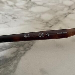 May include: Close-up of the arm of a pair of sunglasses with a brown tortoiseshell pattern. The Ray-Ban logo is visible, along with the text "T", "CE", "UK CA", and "MADE IN CHINA".