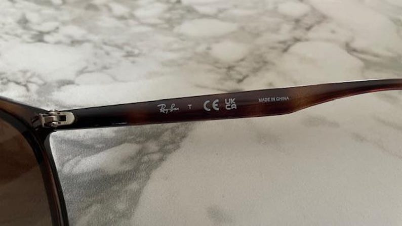 May include: Close-up of the arm of a pair of sunglasses with a brown tortoiseshell pattern. The Ray-Ban logo is visible, along with the text "T", "CE", "UK CA", and "MADE IN CHINA".