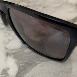 May include: Close-up of black sunglasses with dark lenses. The sunglasses have a rectangular frame and are resting on a light-colored surface. The lenses reflect the surrounding environment.
