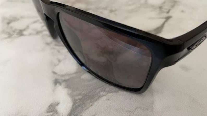May include: Close-up of black sunglasses with dark lenses. The sunglasses have a rectangular frame and are resting on a light-colored surface. The lenses reflect the surrounding environment.