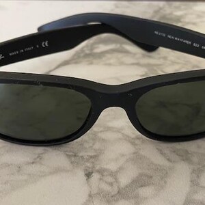May include: Black Ray-Ban Wayfarer sunglasses with green-tinted lenses. The sunglasses have a matte finish and the Ray-Ban logo is visible on the lens. The arms of the sunglasses are folded open.