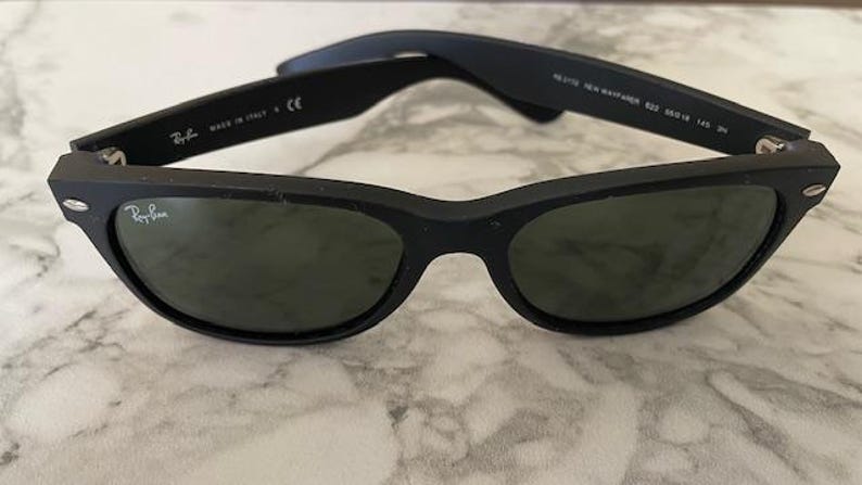 May include: Black Ray-Ban Wayfarer sunglasses with green-tinted lenses. The sunglasses have a matte finish and the Ray-Ban logo is visible on the lens. The arms of the sunglasses are folded open.
