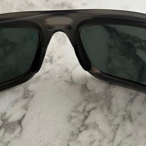 May include: Close-up of dark grey sunglasses with dark green lenses. The sunglasses have a rectangular shape and a bridge over the nose. The lenses are slightly reflective.
