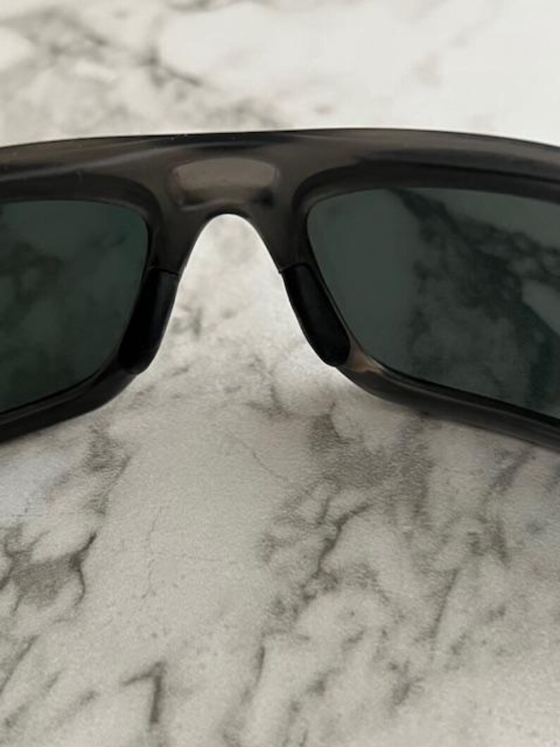 May include: Close-up of dark grey sunglasses with dark green lenses. The sunglasses have a rectangular shape and a bridge over the nose. The lenses are slightly reflective.