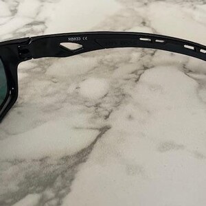 May include: Black sunglasses with a curved arm design. The lenses have a reflective, blue-green tint. The frame is black and features the text "NB832 CE". The sunglasses are shown on a white and gray marble surface.