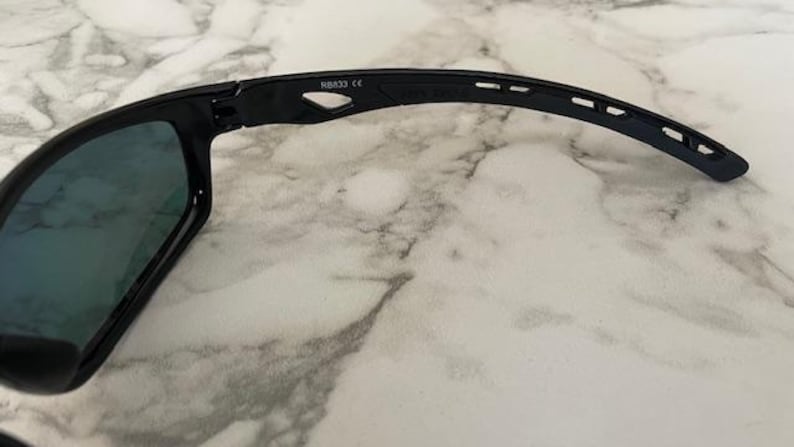 May include: Black sunglasses with a curved arm design. The lenses have a reflective, blue-green tint. The frame is black and features the text "NB832 CE". The sunglasses are shown on a white and gray marble surface.