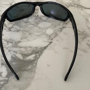 May include: Black sunglasses with a curved frame and dark lenses. The sunglasses are designed for sports or outdoor activities. The arms of the sunglasses curve down and away from the lenses.