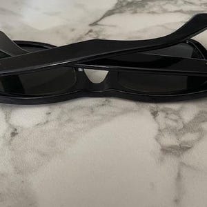 K&ouml;nnte beinhalten: Schwarze Sonnenbrille mit klassischem Design, dunklen Gl&auml;sern und dicken, rechteckigen Rahmen. Die B&uuml;gel sind &uuml;ber den Gl&auml;sern zusammengeklappt. Das Ray-Ban-Logo ist auf den B&uuml;geln sichtbar. Die Sonnenbrille liegt auf einer Marmoroberfl&auml;che.