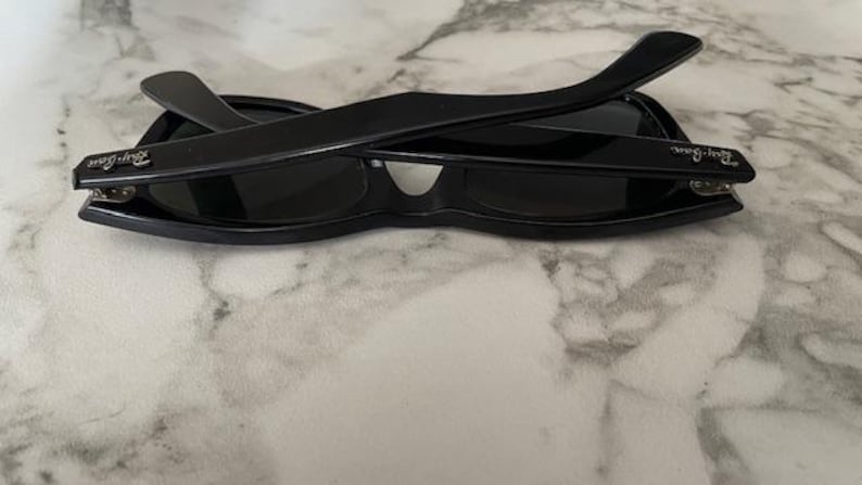 K&ouml;nnte beinhalten: Schwarze Sonnenbrille mit klassischem Design, dunklen Gl&auml;sern und dicken, rechteckigen Rahmen. Die B&uuml;gel sind &uuml;ber den Gl&auml;sern zusammengeklappt. Das Ray-Ban-Logo ist auf den B&uuml;geln sichtbar. Die Sonnenbrille liegt auf einer Marmoroberfl&auml;che.