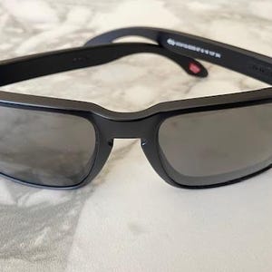 May include: Black sunglasses with a rectangular frame and gray lenses. The sunglasses have a matte finish and small silver rivets on the corners of the frame. The arms of the sunglasses are black and have text printed on them.