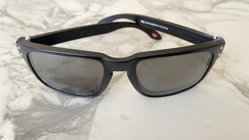 May include: Black sunglasses with a rectangular frame and gray lenses. The sunglasses have a matte finish and small silver rivets on the corners of the frame. The arms of the sunglasses are black and have text printed on them.
