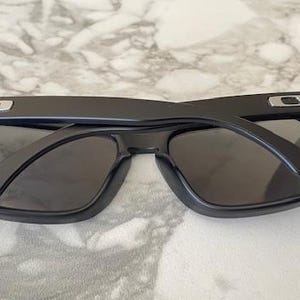 May include: Black sunglasses with a rectangular frame and dark lenses. The sunglasses have a matte finish and a silver accent on the temples. The arms are folded open, revealing the interior of the frame.