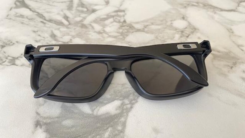 May include: Black sunglasses with a rectangular frame and dark lenses. The sunglasses have a matte finish and a silver accent on the temples. The arms are folded open, revealing the interior of the frame.