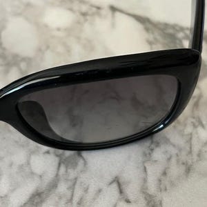 K&ouml;nnte beinhalten: Schwarze Cat-Eye-Sonnenbrille mit dunkel get&ouml;nten Gl&auml;sern. Die Sonnenbrille hat ein schlankes, modernes Design mit gl&auml;nzendem Finish. Der Rahmen ist dick und kantig.