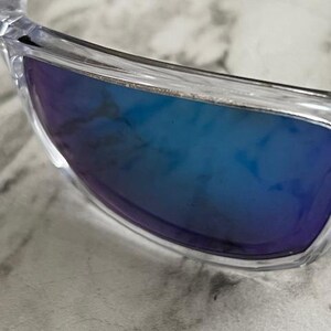 May include: Clear-framed sunglasses with blue gradient lenses. The lenses transition from a deep blue to a lighter shade, offering a stylish and modern look. The close-up shot highlights the sleek design and the quality of the materials.
