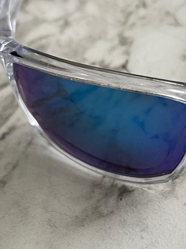 May include: Clear-framed sunglasses with blue gradient lenses. The lenses transition from a deep blue to a lighter shade, offering a stylish and modern look. The close-up shot highlights the sleek design and the quality of the materials.