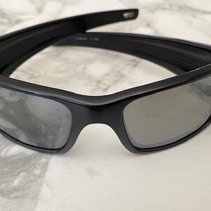 May include: Black sunglasses with a rectangular frame and grey lenses. The sunglasses are folded open, showcasing the sleek design. The lenses are reflective, and the frame appears to be made of a durable material.