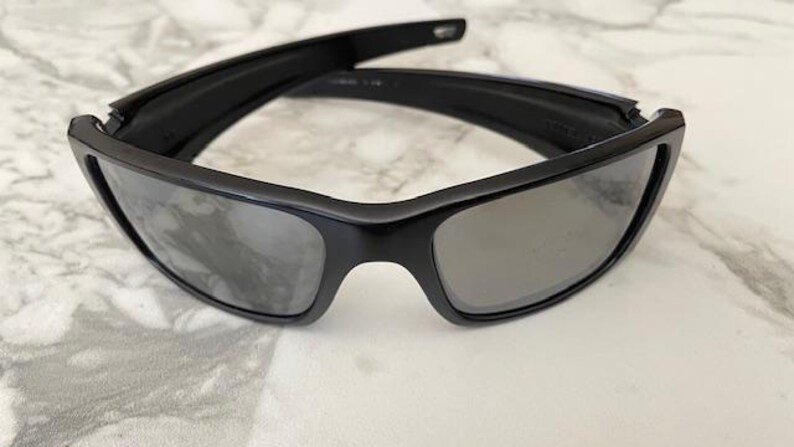 May include: Black sunglasses with a rectangular frame and grey lenses. The sunglasses are folded open, showcasing the sleek design. The lenses are reflective, and the frame appears to be made of a durable material.