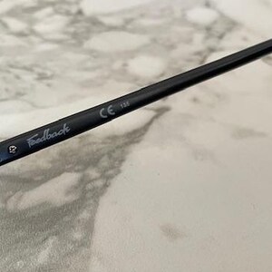 May include: Close-up of black sunglasses with a round lens shape. The arm of the glasses has the word "Feedback" printed in a light color, along with the CE marking and the number 133.