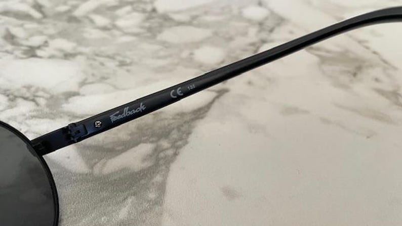 May include: Close-up of black sunglasses with a round lens shape. The arm of the glasses has the word "Feedback" printed in a light color, along with the CE marking and the number 133.