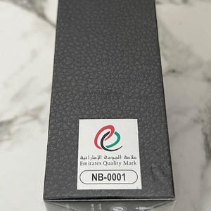 May include: A rectangular black box with a textured surface. A white sticker displays the Emirates Quality Mark logo and the text "NB-0001". The box is likely packaging for a product.