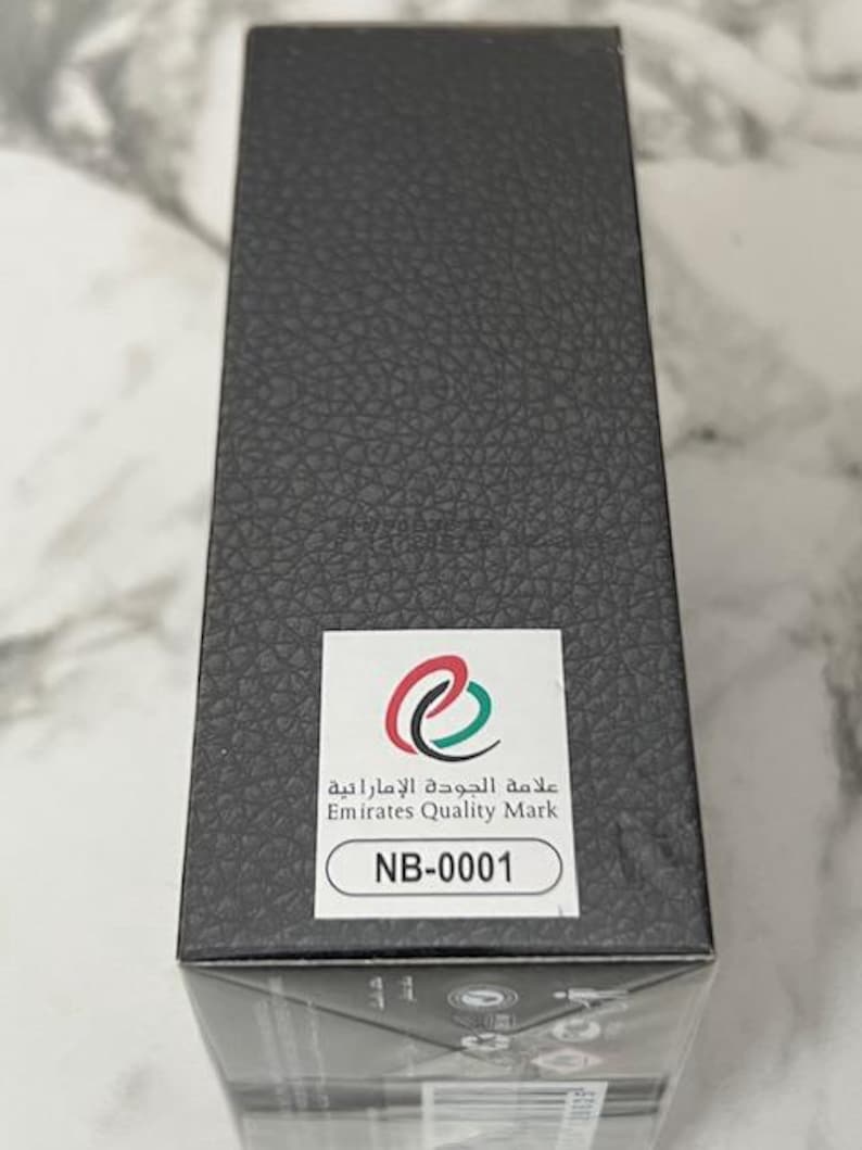 May include: A rectangular black box with a textured surface. A white sticker displays the Emirates Quality Mark logo and the text "NB-0001". The box is likely packaging for a product.