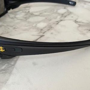 May include: Black sunglasses with a gold anchor emblem on the frame. The sunglasses have a sleek, modern design with dark lenses and curved temples. The frame is made of a matte black material.