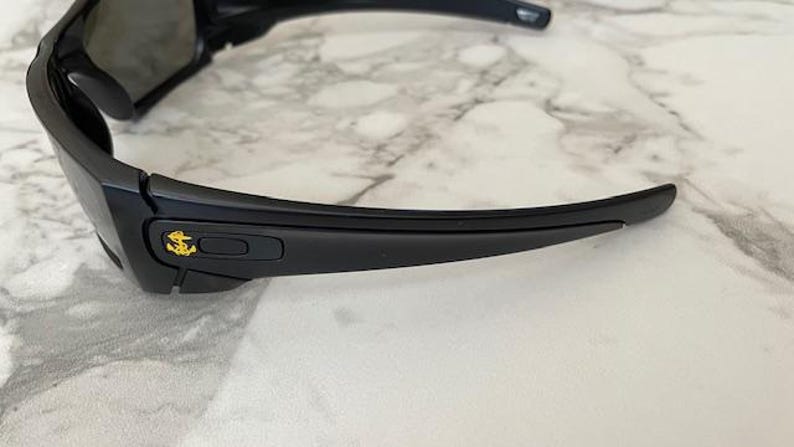 May include: Black sunglasses with a gold anchor emblem on the frame. The sunglasses have a sleek, modern design with dark lenses and curved temples. The frame is made of a matte black material.