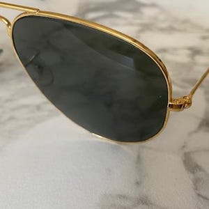May include: Gold-rimmed aviator sunglasses with dark lenses. The sunglasses have a classic teardrop shape and a thin gold frame. The lenses are a dark, solid color, and the temples are thin and gold.