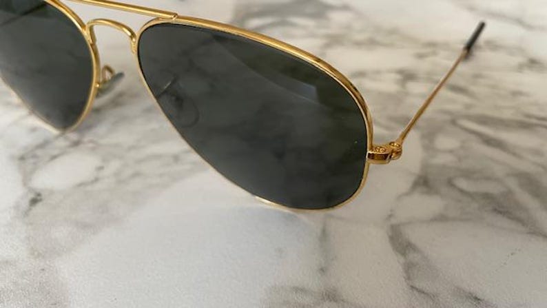 May include: Gold-rimmed aviator sunglasses with dark lenses. The sunglasses have a classic teardrop shape and a thin gold frame. The lenses are a dark, solid color, and the temples are thin and gold.