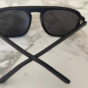 Puede incluir: Gafas de sol negro mate con una barra superior gruesa que conecta las dos lentes tintadas oscuras. Las patillas est&aacute;n cruzadas en la parte delantera, mostrando un acabado negro liso y s&oacute;lido en los marcos y brazos, con un aspecto uniforme.
