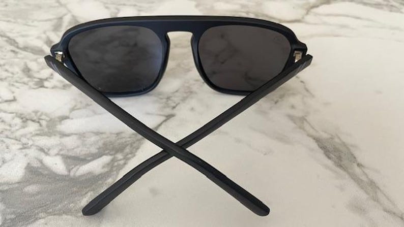 Puede incluir: Gafas de sol negro mate con una barra superior gruesa que conecta las dos lentes tintadas oscuras. Las patillas est&aacute;n cruzadas en la parte delantera, mostrando un acabado negro liso y s&oacute;lido en los marcos y brazos, con un aspecto uniforme.