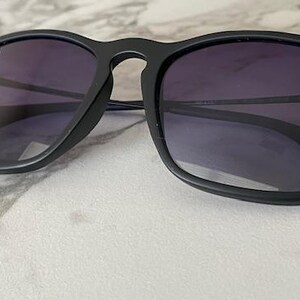 May include: Black sunglasses with dark purple lenses. The sunglasses have a square frame and a matte finish. The Ray-Ban logo is visible on the upper left lens. The sunglasses are resting on a marble surface.