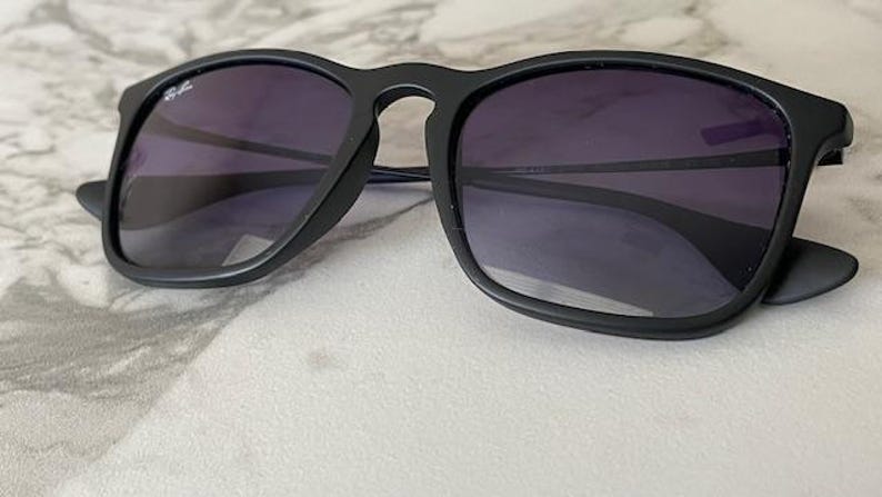 May include: Black sunglasses with dark purple lenses. The sunglasses have a square frame and a matte finish. The Ray-Ban logo is visible on the upper left lens. The sunglasses are resting on a marble surface.