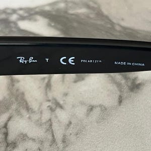 K&ouml;nnte beinhalten: Nahaufnahme des B&uuml;gels einer schwarzen Sonnenbrille. Das Ray-Ban-Logo ist sichtbar, zusammen mit den Texten "T", "CE", "POLARIZED" und "MADE IN CHINA". Die Sonnenbrille liegt auf einer hellen Oberfl&auml;che.
