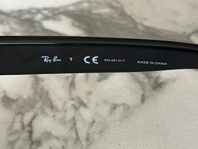 K&ouml;nnte beinhalten: Nahaufnahme des B&uuml;gels einer schwarzen Sonnenbrille. Das Ray-Ban-Logo ist sichtbar, zusammen mit den Texten "T", "CE", "POLARIZED" und "MADE IN CHINA". Die Sonnenbrille liegt auf einer hellen Oberfl&auml;che.