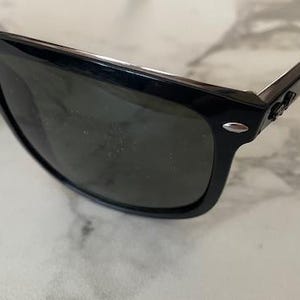 Puede incluir: Gafas de sol negras con lentes oscuras. Las gafas tienen un dise&ntilde;o de montura rectangular cl&aacute;sico. Las patillas presentan un acento plateado cerca de las bisagras. Las lentes son oscuras, proporcionando sombra y protecci&oacute;n solar.